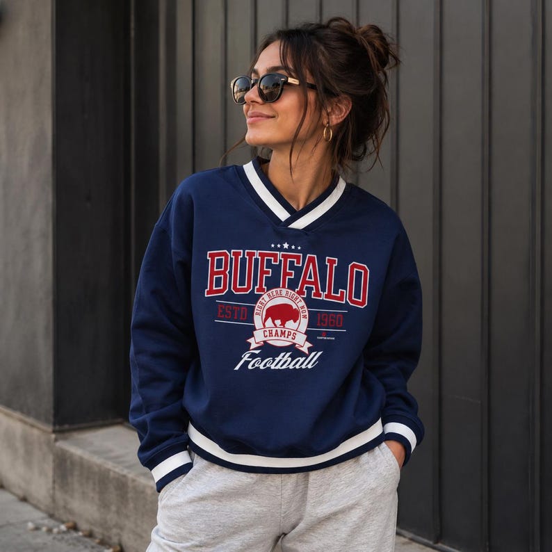 May include: A navy blue long-sleeve sweatshirt with a V-neck collar and white striped trim at the cuffs and hem. The front features red and white text reading BUFFALO, ESTD 1960, CHAMPS, and Football, with a central graphic of a buffalo silhouette.