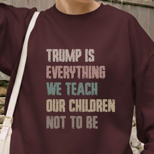 Puede incluir: Sudadera granate con el texto "TRUMP IS EVERYTHING WE TEACH OUR CHILDREN NOT TO BE" en una fuente desgastada. La imagen muestra a una persona que lleva la sudadera.