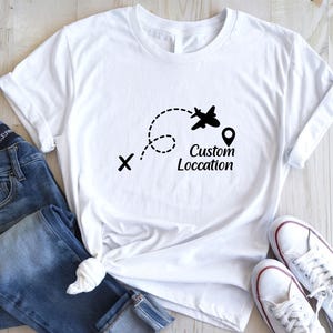 May include: White t-shirt with a black graphic of an airplane following a dotted line, a location pin, and the words "Custom Location". The shirt is paired with blue jeans and white sneakers.