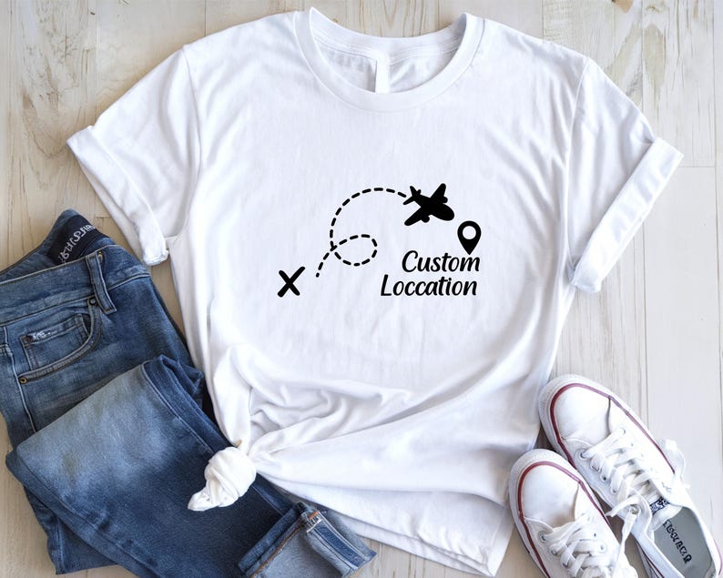May include: White t-shirt with a black graphic of an airplane following a dotted line, a location pin, and the words "Custom Location". The shirt is paired with blue jeans and white sneakers.