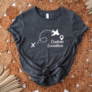 May include: Dark gray t-shirt with a white graphic of an airplane, dotted flight path, and location pin. The text "Custom Location" is below the airplane graphic. The shirt is knotted at the bottom.