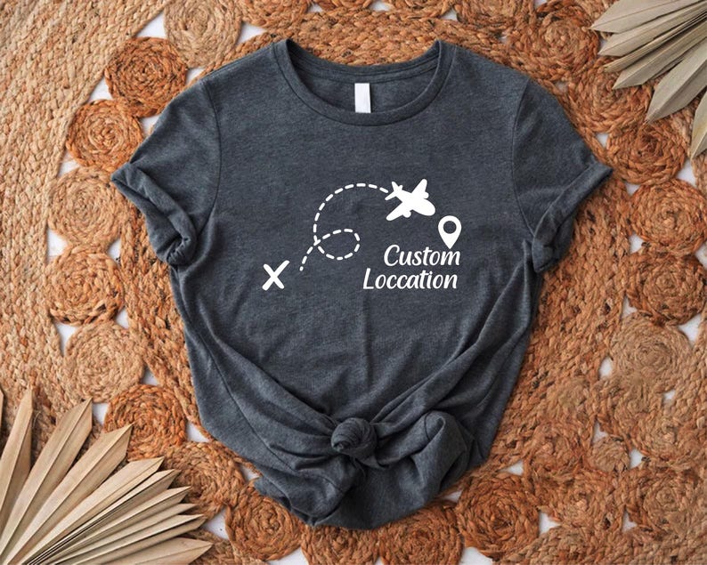 May include: Dark gray t-shirt with a white graphic of an airplane, dotted flight path, and location pin. The text "Custom Location" is below the airplane graphic. The shirt is knotted at the bottom.