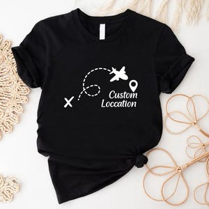 May include: Black t-shirt with a white graphic of an airplane following a dotted line, a location pin, and the words "Custom Location". The shirt is made of a soft material and is suitable for travel enthusiasts.