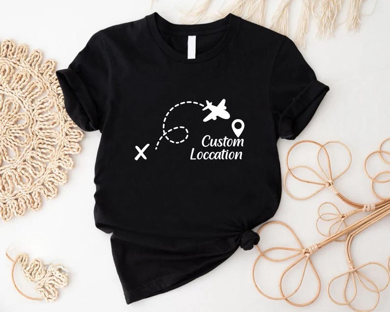 May include: Black t-shirt with a white graphic of an airplane following a dotted line, a location pin, and the words "Custom Location". The shirt is made of a soft material and is suitable for travel enthusiasts.