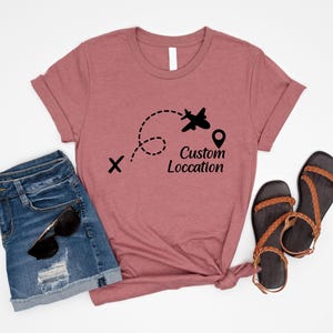 May include: A dusty rose t-shirt with a black airplane graphic and the words "Custom Location". The shirt is paired with denim shorts, sunglasses, and brown sandals, suggesting a travel theme.