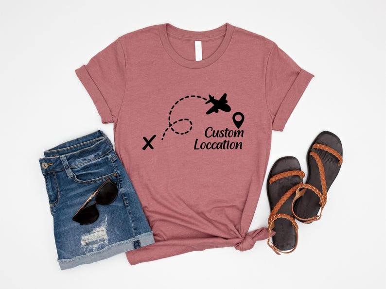 May include: A dusty rose t-shirt with a black airplane graphic and the words "Custom Location". The shirt is paired with denim shorts, sunglasses, and brown sandals, suggesting a travel theme.