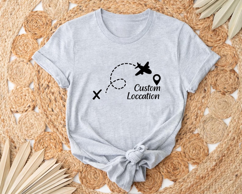 May include: Light gray t-shirt featuring a black graphic of an airplane following a dotted line, with a location pin and the words "Custom Location". The shirt is knotted at the bottom.
