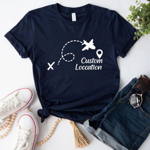 May include: Navy blue t-shirt with a white graphic of an airplane following a dotted line, a location pin, and the text "Custom Location". The shirt is paired with denim shorts, sunglasses, and white sneakers.