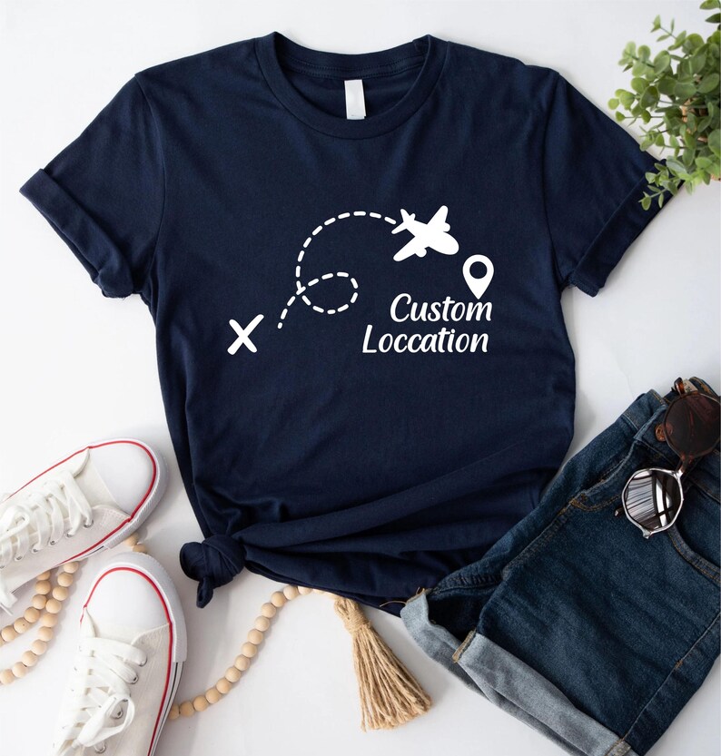 May include: Navy blue t-shirt with a white graphic of an airplane following a dotted line, a location pin, and the text "Custom Location". The shirt is paired with denim shorts, sunglasses, and white sneakers.