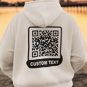 Op de afbeelding: Witte hoodie met een zwart-witte QR-code en de tekst "CUSTOM TEXT" gedrukt op de achterkant.