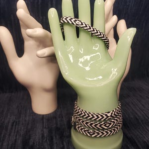 May include: A light green hand-shaped jewelry display holds two black and white woven bracelets. A second white hand-shaped display is in the background. The bracelets have a chevron pattern. The background is a dark blue.