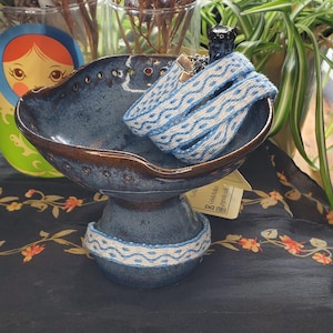 May include: A blue ceramic bowl with a pedestal base, holding a woven blue and white bracelet. The bowl has a dark blue glaze with brown accents. A matching woven band encircles the base. A small, dark-colored object sits atop the bracelet.