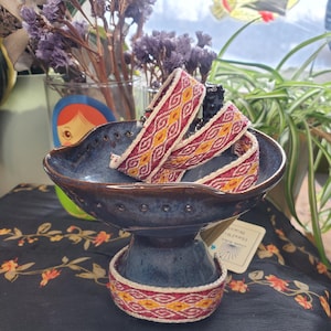 Może przedstawiać: Ciemnoniebieska ceramiczna miska z podstawą na cokole zawiera kilka tkanych bransoletek. Bransoletki mają powtarzający się geometryczny wzór w kolorze czerwonym, żółtym i kremowym. Misa stoi na czarnej tkaninie z kwiatowym wzorem.