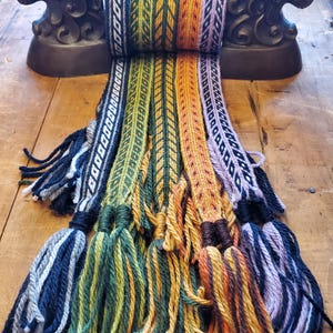 May include: A collection of colorful woven belts with decorative tassels. The belts feature intricate patterns in shades of blue, green, orange, and purple. The tassels are made of thick yarn in coordinating colors. The belts are displayed on a wooden surface.