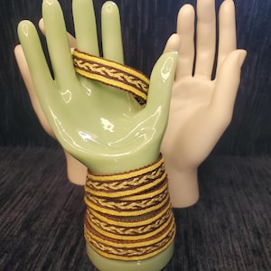 Może przedstawiać: Stos tkanych bransoletek w odcieniach żółci i brązu jest prezentowany na jasnej, zielonej ceramicznej dłoni. Druga ceramiczna dłoń znajduje się w tle. Bransoletki mają pleciony wzór.