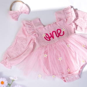 Pode incluir: Um conjunto de roupa de bebé rosa com uma faixa de cabelo. O conjunto apresenta mangas compridas, uma saia de tule e a palavra "one" bordada no corpete. Pequenos detalhes de flores brancas adornam a saia. A faixa tem um laço a condizer.