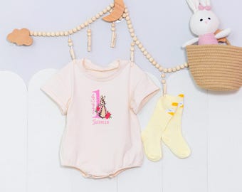 Könnte beinhalten: Ein rosa Baby-Strampler mit einem Peter-Rabbit-Motiv und dem Text "my first cake" und "Jasmin". Der Strampler hängt an einer Wäscheleine aus Holz mit einem Holzmond und einer Holzwolke.