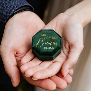May include: A dark green velvet octagonal ring box featuring gold lettering that reads MR. & MRS. Brouns and the date 1.9.2025. The box has a smooth, plush texture and a geometric shape, designed to hold small jewelry items securely.