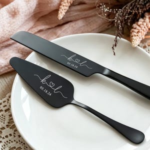 Puede incluir: Un juego de cuchillo y pala para pastel en color negro mate sobre un plato blanco. Ambas piezas presentan un grabado blanco con las letras K y L unidas por un corazón, seguido de la fecha 05.18.24 en una tipografía cursiva y elegante.