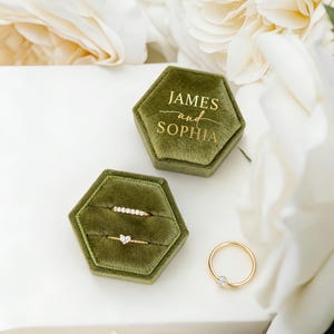 以下が含まれることがあります： 「JAMES and SOPHIA」の金色の文字が入った、オリーブグリーンのベルベットのリングボックス。2つのゴールドリングが2つ目のボックスに、3つ目のリングがボックスの隣に展示されています。背景には白い花があります。
