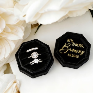 Może przedstawiać: Trzy srebrne pierścionki w czarnym aksamitnym pudełku na pierścionki, z drugim pudełkiem z napisem "MR. & MRS. Browns 1.9.2025" złotymi literami. Pudełka otoczone są białymi kwiatami.