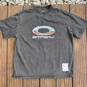 Pode incluir: Uma t-shirt de manga curta cinzento-carvão com uma textura desgastada e esburacada em toda a peça. O peito apresenta um grande gráfico oval em azul claro e laranja com o texto "SATISFY" por baixo. Uma pequena etiqueta branca está fixada.