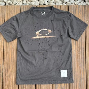 Pode incluir: Uma t-shirt de manga curta em cinzento-carvão com um acabamento desgastado e pequenos orifícios por toda a superfície. A parte frontal apresenta um gráfico de uma estrutura oval com linhas verticais. A etiqueta interior indica Oakley Satisfy.