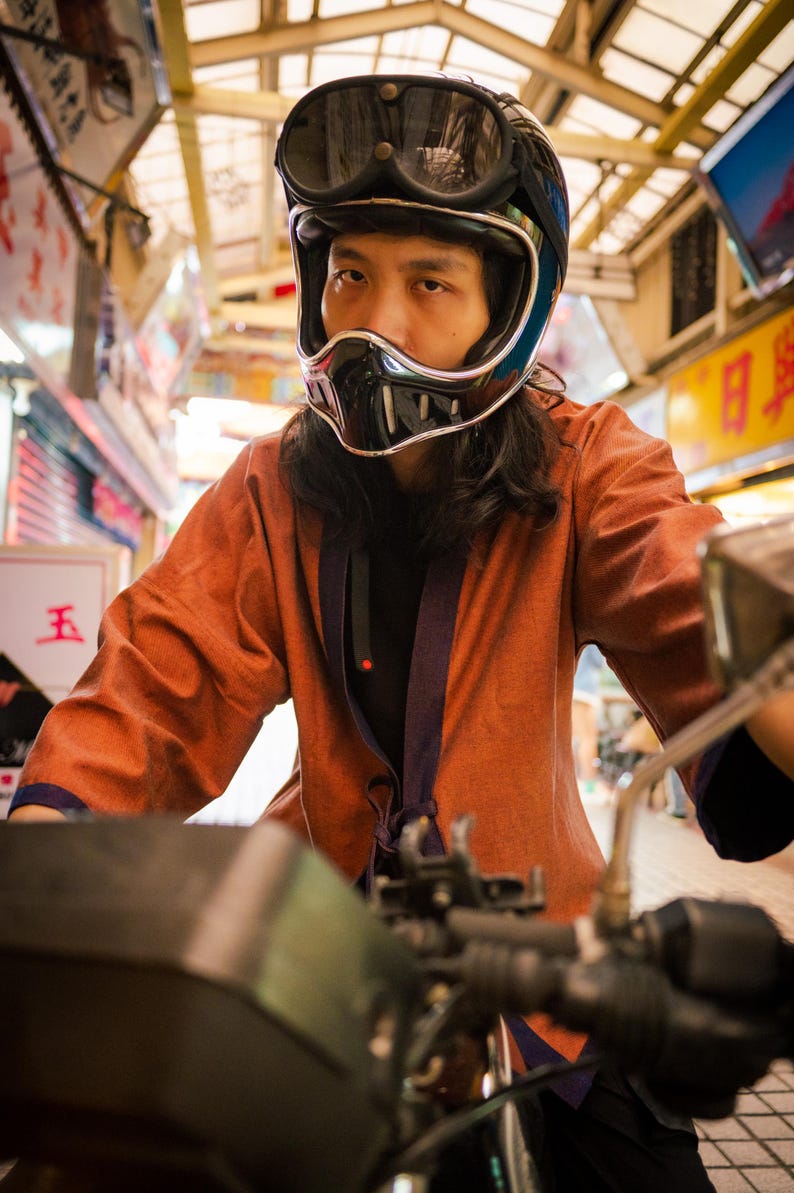 May include: A person wearing a black motorcycle helmet with a visor and a brown jacket. The person is riding a motorcycle.