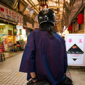 May include: A person wearing a black helmet and a dark blue shirt sits on a black motorcycle. The person is facing away from the camera. The motorcycle is parked in a covered market area.