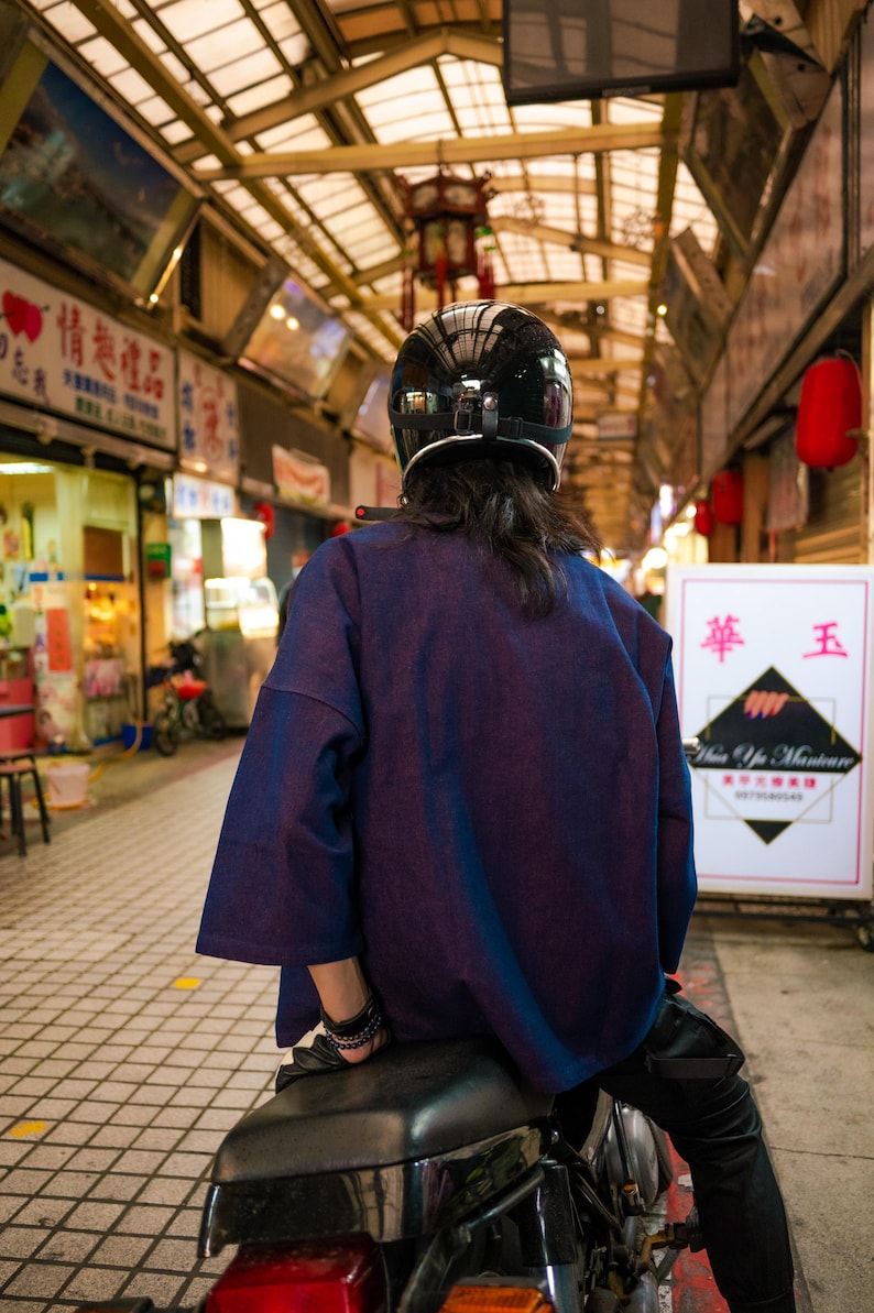 May include: A person wearing a black helmet and a dark blue shirt sits on a black motorcycle. The person is facing away from the camera. The motorcycle is parked in a covered market area.