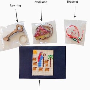 May include: An assortment of handmade items, including a wooden key ring with the text "Will Return", a necklace with a red and gold pendant, and a red and green bracelet. A postcard with a cross-stitch design is also included.