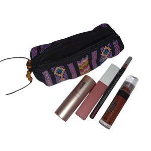 May include: A black and purple striped fabric makeup bag with a zipper closure and wooden beads. The bag is open and contains a pink lipstick, a nude lipstick, a black eyeliner pencil, and a brown lip gloss.