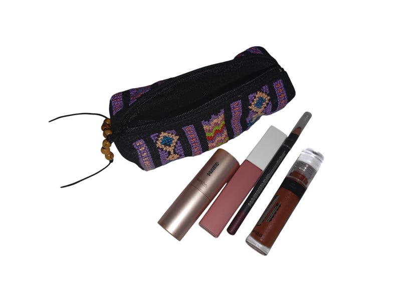 May include: A black and purple striped fabric makeup bag with a zipper closure and wooden beads. The bag is open and contains a pink lipstick, a nude lipstick, a black eyeliner pencil, and a brown lip gloss.