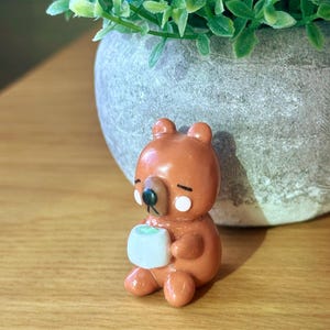 May include: A brown bear figurine holding a small, square, light blue object. The bear has white accents around the eyes and a black nose. The figurine is sitting on a wooden surface, with a potted plant in the background.