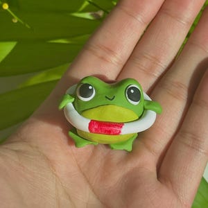 Pode incluir: Uma figura de sapo verde usando um salva-vidas branco e vermelho. O sapo tem olhos grandes e pretos e um pequeno sorriso.