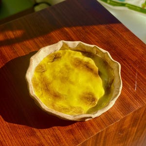 Puede incluir: Un plato de cerámica amarillo con un borde ondulado. El plato tiene un aspecto rústico y hecho a mano y está esmaltado en un color amarillo brillante. El esmalte tiene una textura ligeramente irregular, lo que le da al plato un carácter único.
