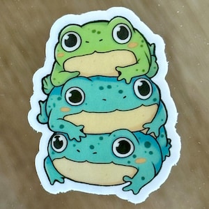 Puede incluir: Una pegatina con tres ranas de dibujos animados apiladas una encima de la otra. La rana de arriba es verde, la rana del medio es azul con manchas blancas y la rana de abajo es azul con manchas blancas. Las tres ranas tienen ojos grandes y sonríen.