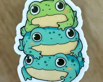 May include: A sticker featuring three cartoon frogs stacked on top of each other. The top frog is green, the middle frog is blue with white spots, and the bottom frog is blue with white spots. All three frogs have large eyes and are smiling.
