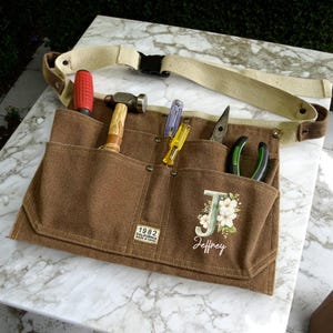 以下が含まれることがあります： 茶色のキャンバス製ツールベルト。複数のポケット、ベージュの調節可能なベルト、黒いバックルが付いています。赤いドライバー、ハンマー、ペンチなどの工具が入っています。前面には「Jeffrey」の名前と文字「J」がパーソナライズされています。