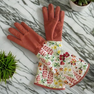 Pu&ograve; includere: Un paio di guanti da giardinaggio lunghi con un palmo in pelle color terracotta e un dorso in cotone con stampa floreale. La stampa floreale presenta fiori rossi, rosa, gialli e verdi su uno sfondo bianco.