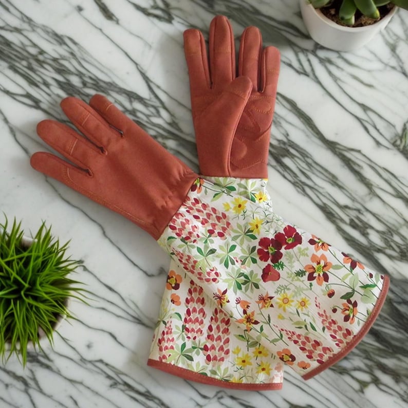 Pu&ograve; includere: Un paio di guanti da giardinaggio lunghi con un palmo in pelle color terracotta e un dorso in cotone con stampa floreale. La stampa floreale presenta fiori rossi, rosa, gialli e verdi su uno sfondo bianco.