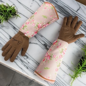 Pu&ograve; includere: Un paio di guanti da giardinaggio marroni con tessuto a stampa floreale che si estende fino alle braccia. I guanti hanno un palmo e dita in pelle marrone, e il tessuto &egrave; rosa con un motivo floreale. I guanti sono etichettati "Badger".