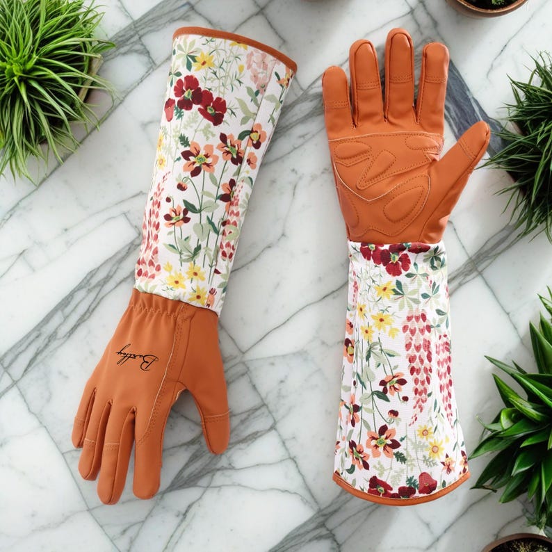 Pu&ograve; includere: Un paio di guanti da giardinaggio lunghi con stampa floreale. I guanti sono in pelle marrone con sfondo bianco e un motivo floreale in tonalit&agrave; di rosso, giallo e rosa. I guanti hanno un polsino lungo che si estende fino al gomito. I guanti sono del marchio "Buddy".