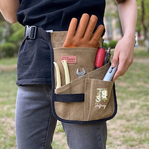 May include: A brown canvas tool belt with multiple pockets, holding tools and orange work gloves. The belt has the text "FLY DADDY" and a floral initial "J" with the name "Jeffrey".