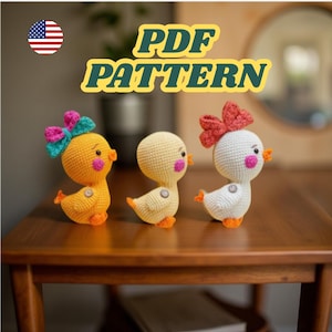 May include: Three crocheted ducklings in yellow and white, each with a bow. The ducklings have orange beaks and feet, and pink cheeks. The text "PDF PATTERN" is displayed in yellow and green.