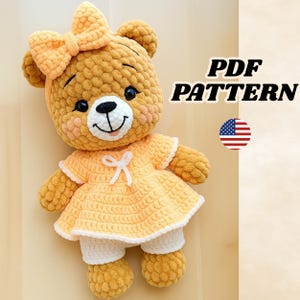 May include: A crocheted teddy bear wearing a yellow dress and bow. The bear has a white muzzle, black eyes, and a smiling mouth. The text "PDF PATTERN" is displayed on the right side of the image, along with the American flag.