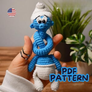 May include: A handmade blue crochet Smurf doll with a white hat and diaper. The doll has black eyes and a smiling expression. The text "PDF PATTERN" is visible in the image. The doll is held in a hand.