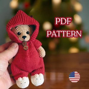 Könnte beinhalten: Ein gehäkelter Teddybär in einem roten Kapuzen-Overall mit weißen Babyschuhen. Der Bär hat ein beiges Gesicht und ist mit zwei Holzknöpfen zugeknöpft. Der Text "PDF PATTERN" ist rot dargestellt. Eine amerikanische Flagge befindet sich in der unteren rechten Ecke.