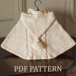 Op de afbeelding: Crèmekleurige gebreide cape met een kabelgebreid ontwerp en een kwast. De cape wordt op een hanger getoond. De woorden "PDF PATTERN" staan onderaan de afbeelding.