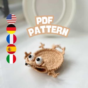 May include: A crocheted brown frog-like coaster with large white and black eyes. The coaster has a circular body and four small legs. The text "PDF PATTERN" is displayed in a light brown font.